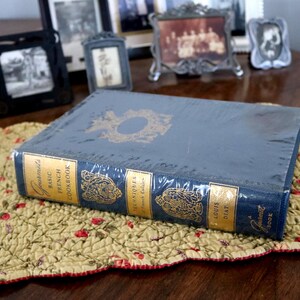May include: A blue hardcover book titled Gourmet's Basic French Cookbook by Louis Diat. The spine features gold lettering and decorative emblems, with a matching gold emblem on the front cover. The book is wrapped in clear plastic protective film.