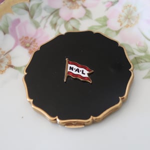 May include: A black and gold compact with a white and red flag with the letters "N-A-L" on it.