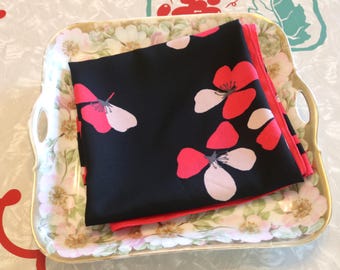 May include: A black floral scarf with pink and white flower designs is neatly folded on a decorative tray. The tray has a floral pattern with gold trim and handles. The scarf's edges are a bright red.