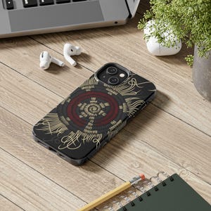 May include: Black phone case with a circular design in red, gold, and black. The case features a detailed pattern resembling a sun or mandala. The phone case is on a wooden surface, with a laptop, earbuds, and a notebook in the background.