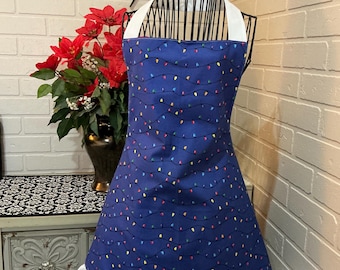 May include: A blue apron with a pattern of colorful string lights. The apron has a white halter top and a white ruffled hem. The word "sew" is displayed in black cursive on the wall behind the apron.