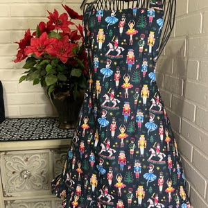 May include: A dark blue apron with a festive Nutcracker print, featuring ballerinas, toy soldiers, and rocking horses. The apron has a ruffled hem and is displayed on a mannequin. The word "sew" is visible in the background.