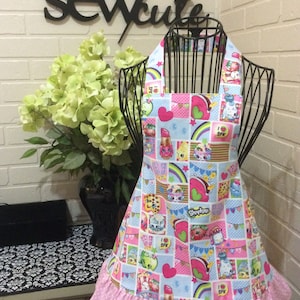 May include: A pink and blue children's apron with a colorful print of cartoon characters and the words "Shopkins" and "I Spy". The apron has a pink ruffle trim.