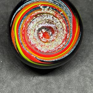 May include: A spherical glass object featuring a swirling vortex pattern of red, yellow, and blue colors. The center of the sphere contains a dense cluster of shimmering, reflective silver-toned glitter particles embedded within the clear glass.