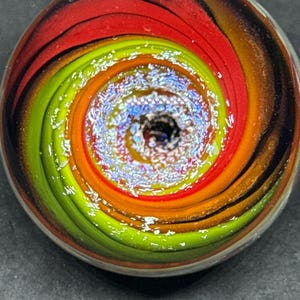 May include: A circular glass marble featuring a swirling vortex pattern of vibrant lime green, bright orange, and deep red colors. The center of the marble contains a textured, iridescent, and glittery core that reflects light with a shimmering effect.