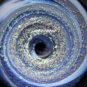 May include: Close-up of a glass art piece with a spiral design. The piece features shades of blue, white, and silver, creating a galaxy-like effect. The center has a dark, deep hole. The glass has a textured, glittery appearance.