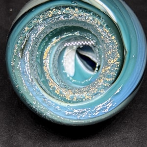 May include: A handcrafted glass paperweight with a swirling, spiral design. The paperweight features shades of teal, blue, and white, with gold flecks embedded within the glass. The spiral center creates a unique visual effect.