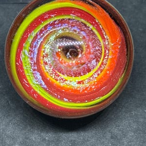 May include: A circular glass object featuring a swirling pattern of vibrant lime green, bright orange, and deep red colors. The surface has a glossy finish with embedded glitter particles that catch the light, creating a spiral effect toward the center.