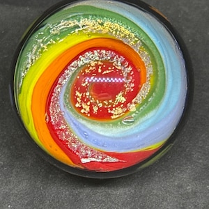 May include: A spherical glass paperweight featuring a swirling vortex pattern of red, orange, yellow, green, and light blue colors. The design incorporates embedded metallic gold and silver flakes throughout the spiral, creating a reflective effect.