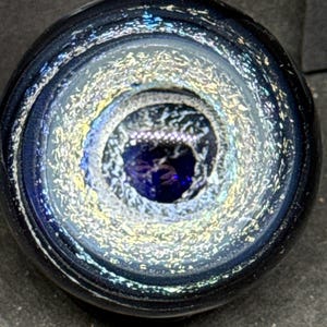 May include: A spherical glass paperweight featuring a deep navy blue core surrounded by a swirling ring of iridescent, shimmering silver and gold flecks. The outer edge of the sphere is a solid, dark translucent blue, creating a layered effect.