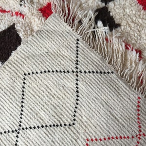 May include: Close-up of a white rug with a black and red diamond pattern. The rug has a fringe border and is woven with a textured surface.