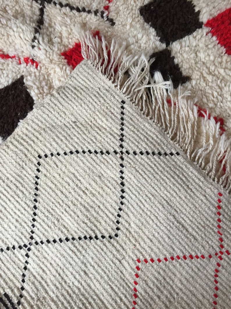 May include: Close-up of a white rug with a black and red diamond pattern. The rug has a fringe border and is woven with a textured surface.