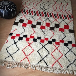 May include: A white, red, and black Moroccan rug with a geometric pattern. The rug has a fringe border.