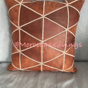 Puede incluir: Una almohada de cuero marr&oacute;n con un patr&oacute;n geom&eacute;trico cosido con hilo blanco. La almohada est&aacute; en un sof&aacute; gris. El texto "@MoroccanGiving" es visible en el centro inferior de la imagen.