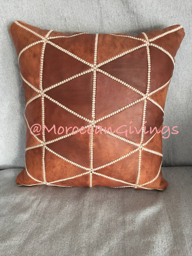 Puede incluir: Una almohada de cuero marr&oacute;n con un patr&oacute;n geom&eacute;trico cosido con hilo blanco. La almohada est&aacute; en un sof&aacute; gris. El texto "@MoroccanGiving" es visible en el centro inferior de la imagen.