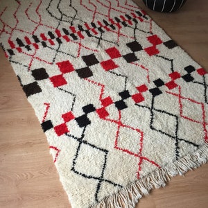 May include: A white, red, and black Moroccan rug with a geometric pattern. The rug has a fringe border and is made of wool.