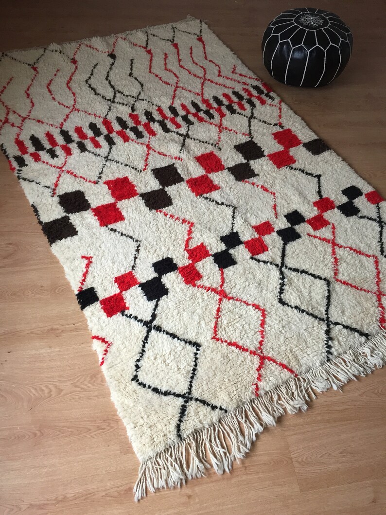 May include: A white, red, and black Moroccan rug with a geometric pattern. The rug has a fringe border and is made of wool.