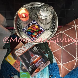 Puede incluir: Una bandeja de plata con dise&ntilde;os intrincados, una vela encendida y una peque&ntilde;a caja de plata con una tapa decorativa. La bandeja est&aacute; sentada sobre una alfombra azul y marr&oacute;n con una almohada de cuero marr&oacute;n con costuras blancas. La almohada tiene un patr&oacute;n geom&eacute;trico.