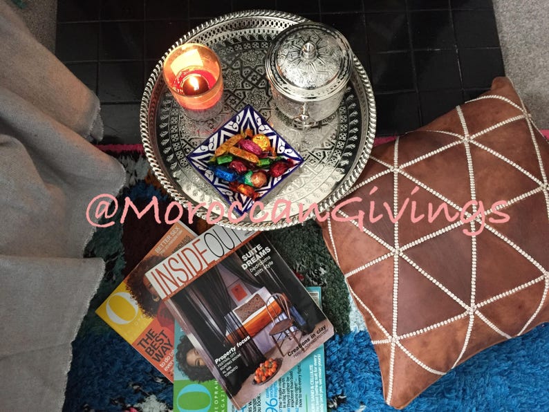 Puede incluir: Una bandeja de plata con dise&ntilde;os intrincados, una vela encendida y una peque&ntilde;a caja de plata con una tapa decorativa. La bandeja est&aacute; sentada sobre una alfombra azul y marr&oacute;n con una almohada de cuero marr&oacute;n con costuras blancas. La almohada tiene un patr&oacute;n geom&eacute;trico.