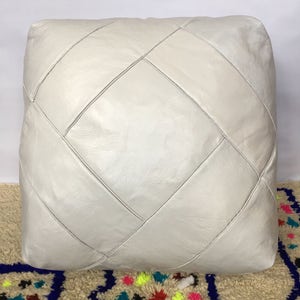 Op de afbeelding: Een witte leren poef met een gestikt patroon in de vorm van een diamant. De poef staat op een tapijt met kleurrijke patronen.