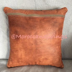 Puede incluir: Una almohada de cuero marr&oacute;n con cierre de cremallera. La almohada tiene un aspecto desgastado y est&aacute; hecha de cuero genuino. La almohada est&aacute; sentada sobre una superficie de tela gris claro. @MoroccanGiving
