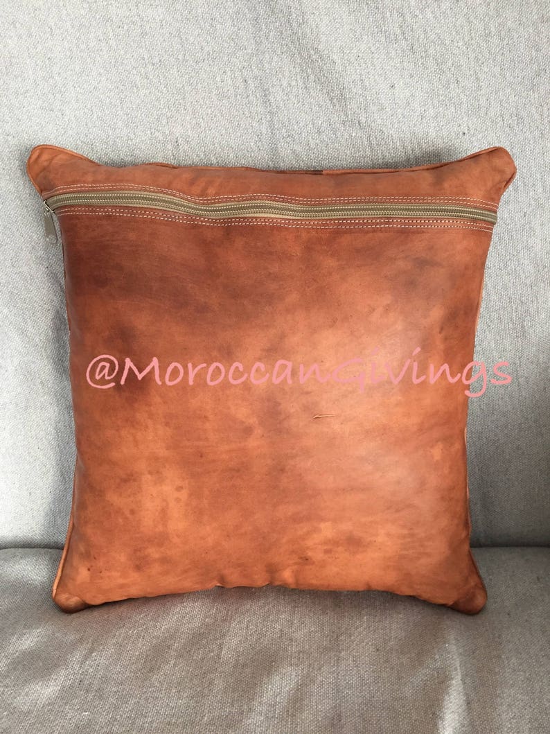 Puede incluir: Una almohada de cuero marr&oacute;n con cierre de cremallera. La almohada tiene un aspecto desgastado y est&aacute; hecha de cuero genuino. La almohada est&aacute; sentada sobre una superficie de tela gris claro. @MoroccanGiving