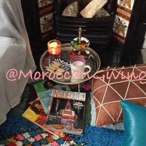 Puede incluir: Una acogedora escena de sala de estar con una chimenea, una manta y una bandeja de galletas y una taza de t&eacute;. La bandeja est&aacute; sobre un soporte decorativo con dise&ntilde;o marroqu&iacute;. Hay revistas en el suelo, incluida una titulada "Inside Out". El texto "@MoroccanGiving" es visible en la imagen.
