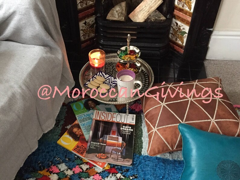 Puede incluir: Una acogedora escena de sala de estar con una chimenea, una manta y una bandeja de galletas y una taza de t&eacute;. La bandeja est&aacute; sobre un soporte decorativo con dise&ntilde;o marroqu&iacute;. Hay revistas en el suelo, incluida una titulada "Inside Out". El texto "@MoroccanGiving" es visible en la imagen.