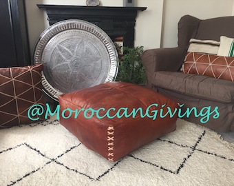 Op de afbeelding: Een bruine leren poef met decoratieve stiksels staat op een wit en zwart gestreept tapijt in een woonkamer. Op de achtergrond zijn een zilveren dienblad en een bruine fauteuil met kussens te zien. De tekst '@MoroccanGiving' is zichtbaar in de afbeelding.