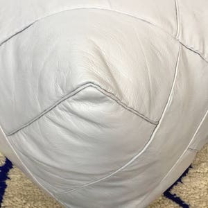 Op de afbeelding: Een witte leren poef met ruitvormige stiksels. De poef staat op een tapijt met een patroon.