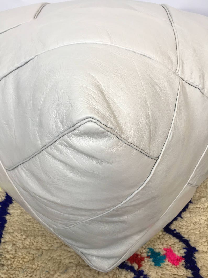 Op de afbeelding: Een witte leren poef met ruitvormige stiksels. De poef staat op een tapijt met een patroon.