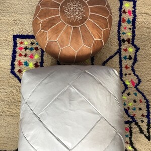 Op de afbeelding: Twee leren poefs, een bruin met een stervormig ontwerp en een wit met een ruitpatroon. De poefs staan op een beige kleed met kleurrijke geometrische patronen.
