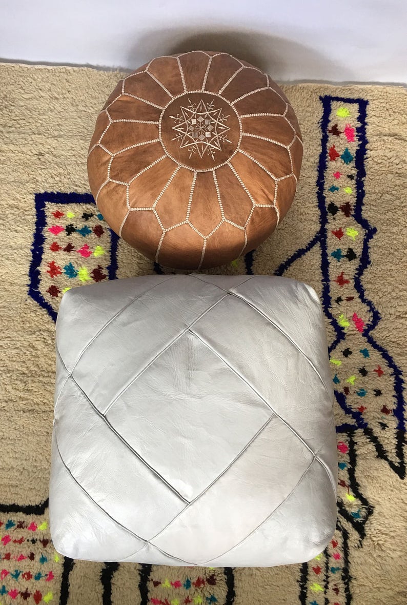Op de afbeelding: Twee leren poefs, een bruin met een stervormig ontwerp en een wit met een ruitpatroon. De poefs staan op een beige kleed met kleurrijke geometrische patronen.