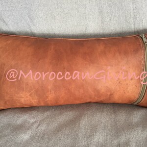 Puede incluir: Una almohada de cuero marr&oacute;n con cierre de cremallera. La almohada tiene un aspecto desgastado y est&aacute; hecha de cuero genuino. La almohada est&aacute; decorada con el texto "@MoroccanGiving" en rosa.