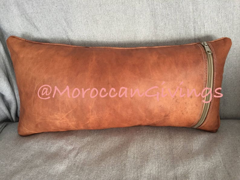 Puede incluir: Una almohada de cuero marr&oacute;n con cierre de cremallera. La almohada tiene un aspecto desgastado y est&aacute; hecha de cuero genuino. La almohada est&aacute; decorada con el texto "@MoroccanGiving" en rosa.
