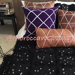 K&ouml;nnte beinhalten: Eine schwarze Wurfdecke mit silbernen Pailletten liegt auf einem Bett mit mehreren Kissen. Die Kissen sind aus braunem Leder mit wei&szlig;en N&auml;hten und ein violettes Kissen mit wei&szlig;en Rauten n&auml;hten. Der Text "@MoroccanGiving" ist im Bild sichtbar.