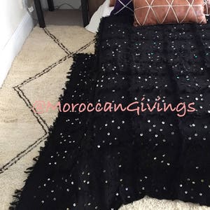 K&ouml;nnte beinhalten: Schwarze Tagesdecke mit Pailletten auf einem wei&szlig;en und braunen gemusterten Teppich. Die Decke hat einen Fransenrand. Der Teppich hat ein Rautenmuster.