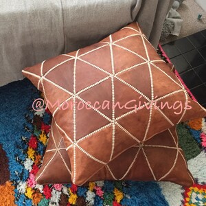 Puede incluir: Dos cojines de cuero marr&oacute;n con un patr&oacute;n geom&eacute;trico y costuras blancas. Los cojines est&aacute;n sobre una alfombra colorida con un fondo azul.