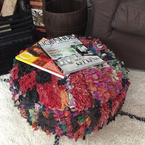 May include: A colourful, round pouf made of fabric scraps, with a magazine titled "Eigenhuis" on top. The pouf is sitting on a white shag rug with black accents.