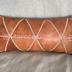Puede incluir: Una almohada de cuero marr&oacute;n con un patr&oacute;n de diamante y diamantes de imitaci&oacute;n blancos alrededor de los bordes. La almohada est&aacute; sobre una superficie de tela gris.