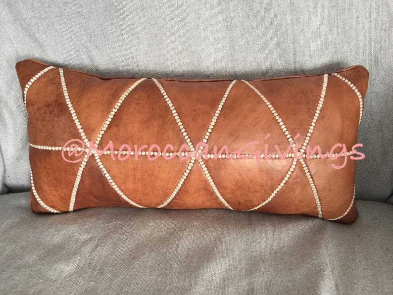 Puede incluir: Una almohada de cuero marr&oacute;n con un patr&oacute;n de diamante y diamantes de imitaci&oacute;n blancos alrededor de los bordes. La almohada est&aacute; sobre una superficie de tela gris.
