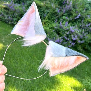 Puede incluir: Una diadema con dos orejas de animal triangulares hechas de fieltro gris en el exterior y piel sintética suave de color rosa claro en el interior. La oreja izquierda está adornada con dos pequeños anillos de metal dorado en el borde superior.