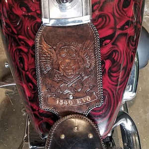 May include: A red motorcycle gas tank with a black and red rose design. The tank has a brown leather patch with a skull and crossbones design and the text "1340 EVO". The motorcycle also has a brown leather seat with a decorative design.