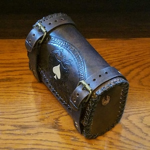 Könnte beinhalten: Eine braune Leder-Motorradsatteltasche mit einem geprägten Design und dem Text "Custom Leather Works" an der Seite. Die Tasche hat einen Schnallenverschluss und einen geflochtenen Lederbesatz.