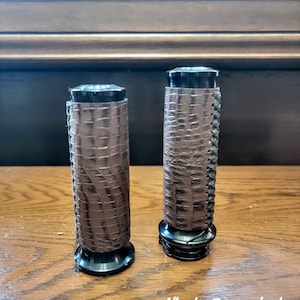 Puede incluir: Dos puños de manillar de motocicleta con revestimiento de cuero marrón con textura de caimán, costuras negras y tapas de metal negro. El texto Alligator Brown coloration y CC Custom Leatherworks es visible abajo a la derecha.