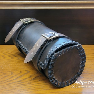 May include: Antique dark brown leather motorcycle saddlebag with two buckles and a braided trim. CC Custom Leatherworks.