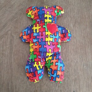 May include: A colorful plush toy bear with a red heart on its chest. The bear is covered in a puzzle piece pattern with various colors, including red, blue, yellow, green, and purple.