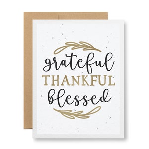 May include: A white card with a gold and black design. The card has a wreath of gold leaves around the words "grateful thankful blessed".