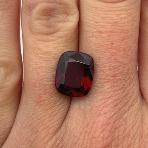 May include: A deep red, cushion-cut gemstone with a faceted surface. The stone displays a dark, saturated crimson hue and a polished finish, reflecting light across its geometric, multi-sided top surface and edges.