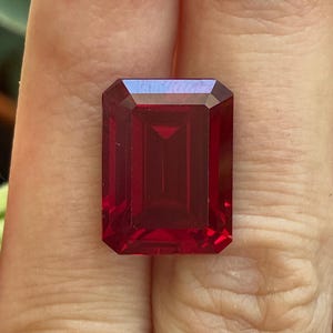 May include: A vibrant, deep red gemstone cut in a classic emerald shape with sharp, clean facets. The stone displays a rich, saturated crimson hue and a high-clarity, transparent surface that reflects light across its rectangular, stepped-cut structure.
